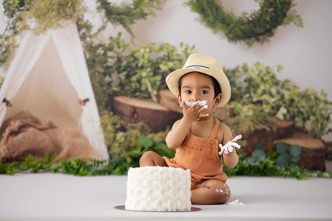 Baby in orange romper and straw hat enjoying cake during a rustic woodland-themed cake smash with greenery and forest backdrop by Cake Smash by Chris in Melbourne.