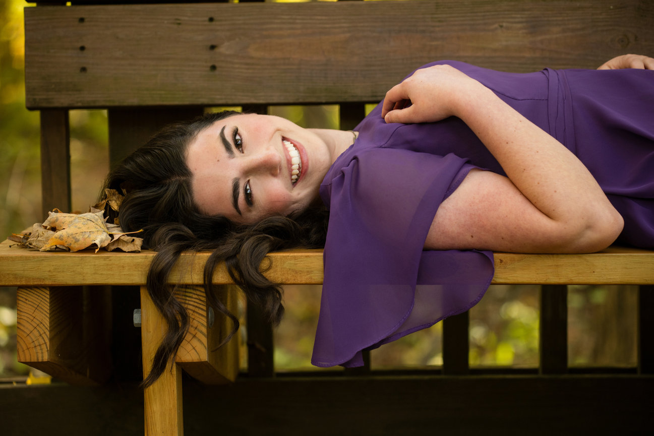 Teen girl in a purple dress lying on a wooden bench, smiling with autumn leaves nearby for senior pictures in Dublin Ohio