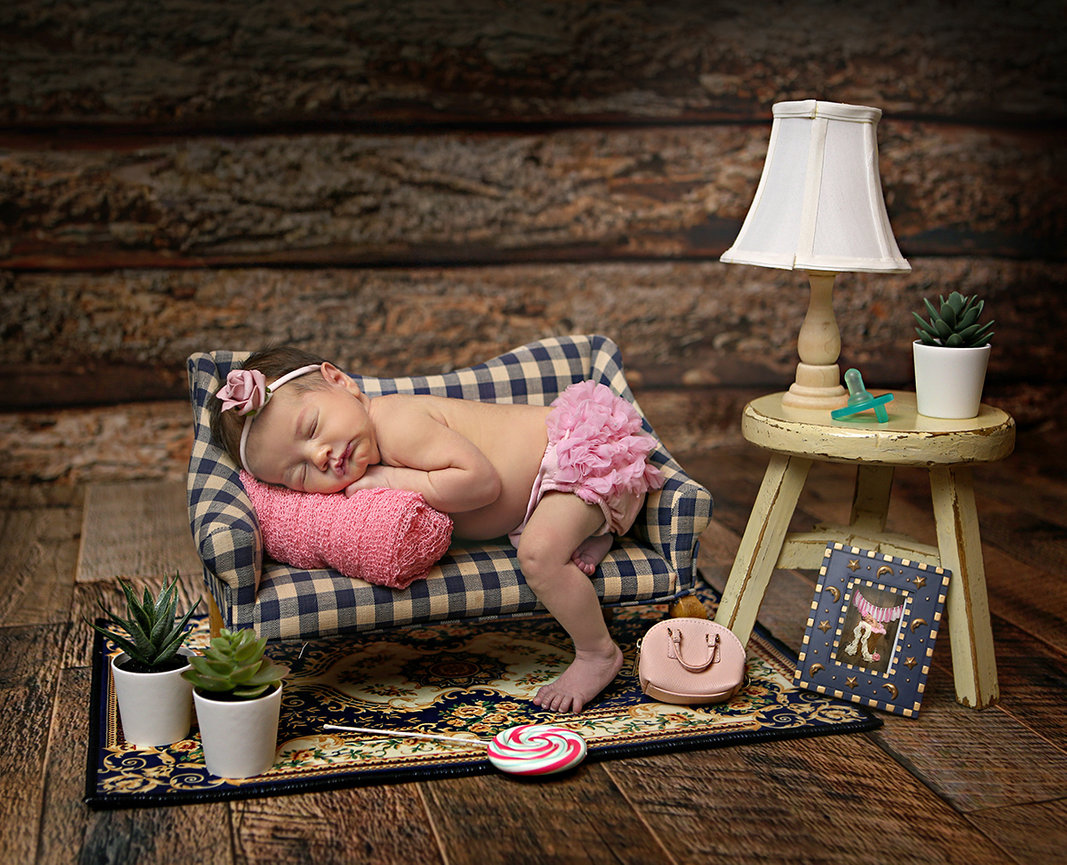 Sleeping baby on a miniature couch with a pink pillow wearing a headband and ruffled shorts beside a small table and lamp.